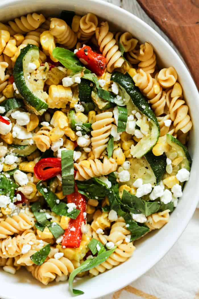 Late summer veggie pasta salad with red peppers, corn, zucchini, fresh basil, and goat cheese, in a white bowl with a wooden spoon