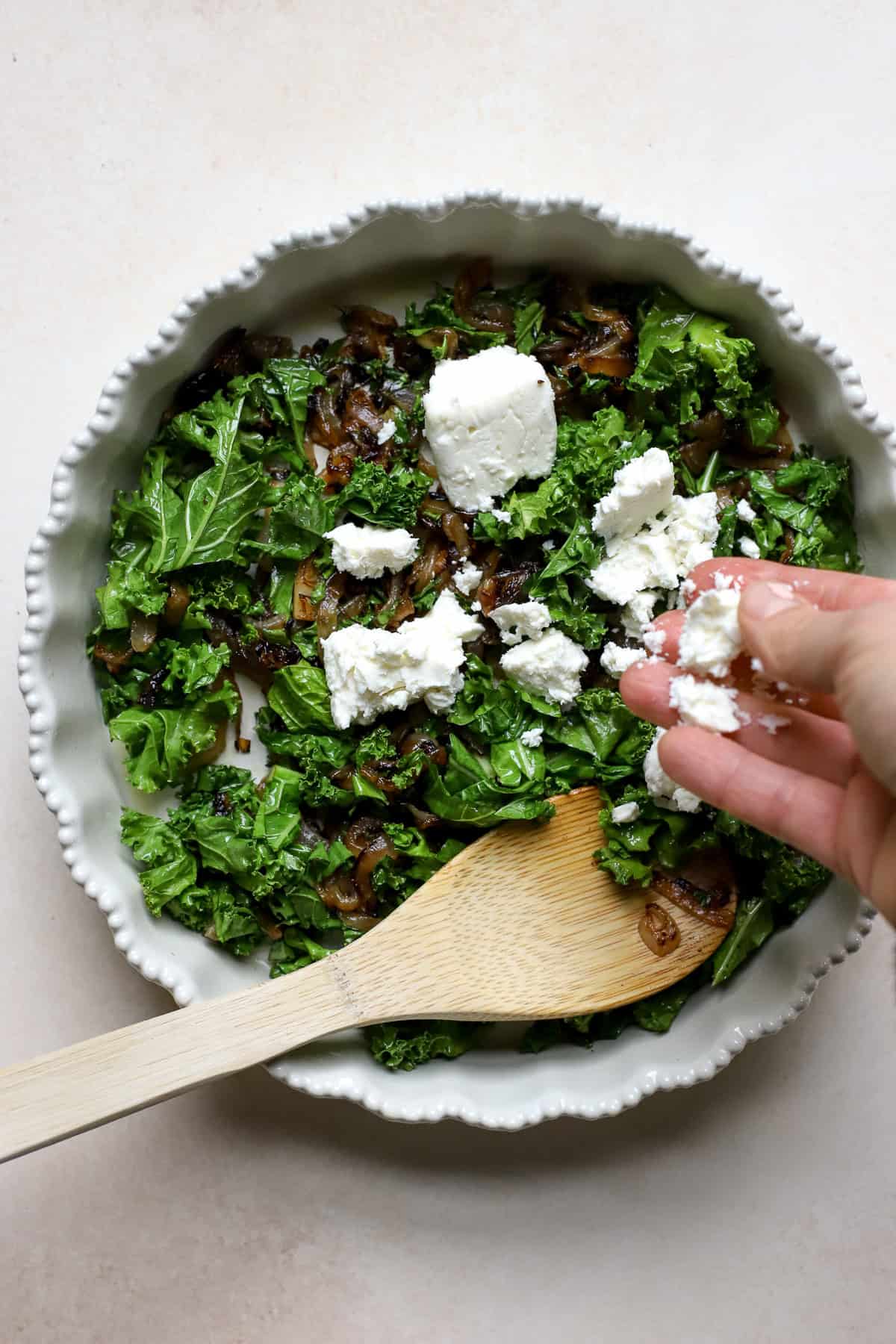 Feta being crumbled into pie dish with kale and caramelized onions.