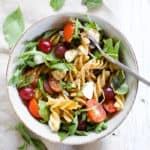 Caprese pasta salad with grapes in a bowl with a silver fork, sitting on a white farmhouse wood surface with a gray linen on the side.