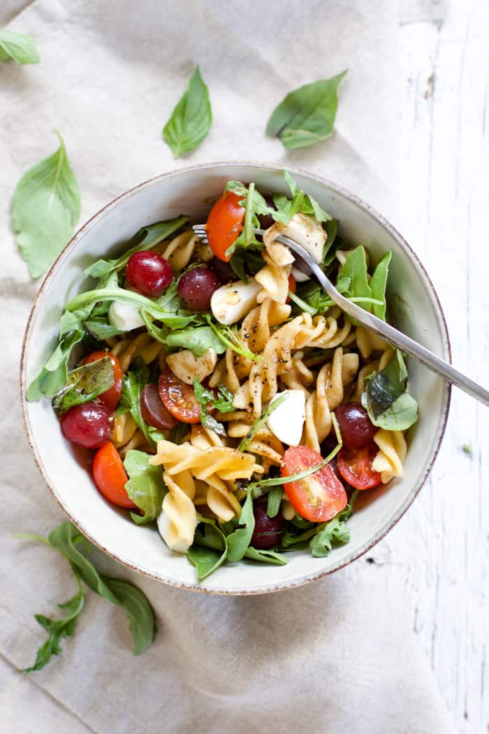 Caprese pasta salad with grapes in a bowl with a silver fork, sitting on a white farmhouse wood surface with a gray linen on the side.