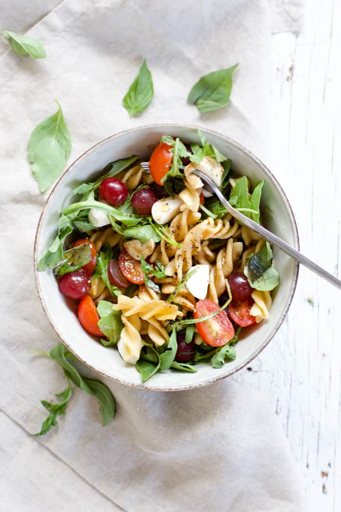 Caprese pasta salad with grapes in a bowl with a silver fork, sitting on a white farmhouse wood surface with a gray linen on the side.