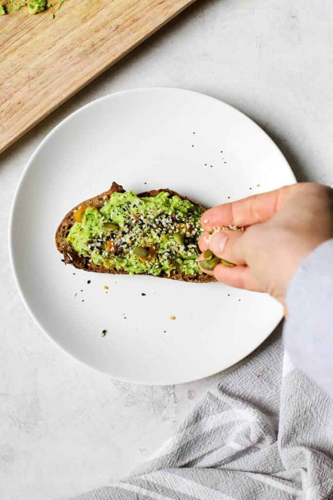 Hand seasoning avocado toast with everything bagel seasoning, hemp seeds, and pepitas.