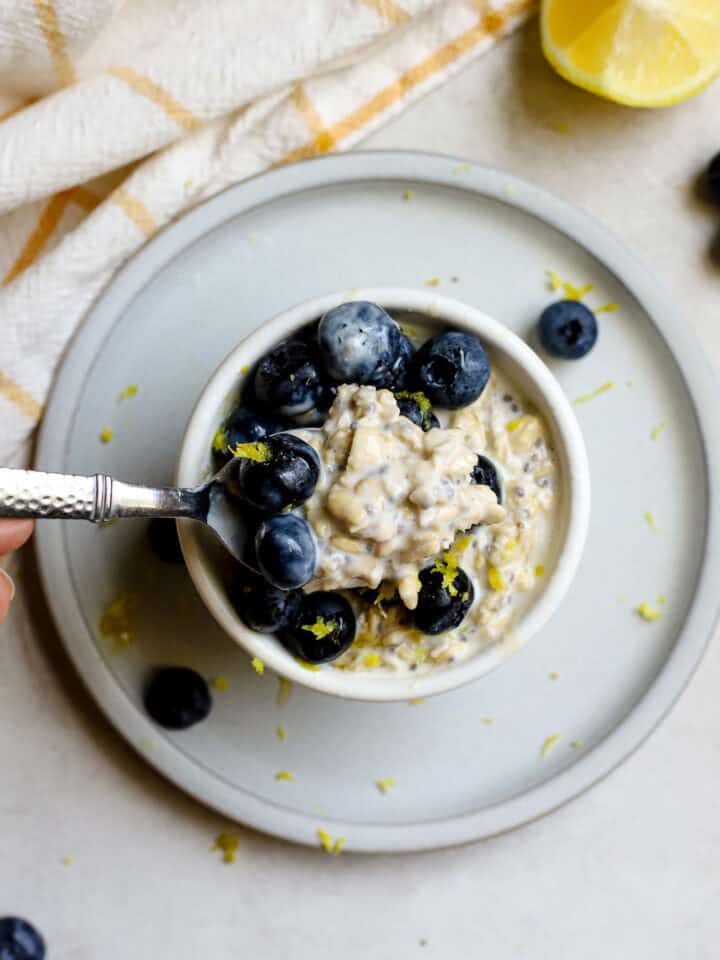 Blueberry lemon overnight oats in small white bowl resting on small blue plate, on gray and white surface. A hand gently starts to scoop a spoonful of oats and blueberries. A few blueberries and a lemon slice sit off to the side with a white and yellow windowpane-printed linen.