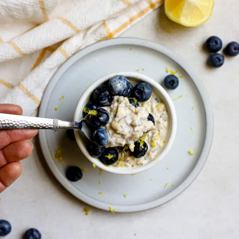 Blueberry lemon overnight oats in small white bowl resting on small blue plate, on gray and white surface. A hand gently starts to scoop a spoonful of oats and blueberries. A few blueberries and a lemon slice sit off to the side with a white and yellow windowpane-printed linen.