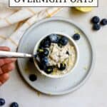 Blueberry lemon overnight oats in small white bowl resting on small blue plate, on gray and white surface. A hand gently starts to scoop a spoonful of oats and blueberries. A few blueberries and a lemon slice sit off to the side with a white and yellow windowpane-printed linen.