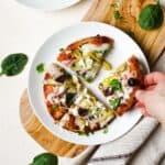 Greek pita pizza slices on white plate with hand picking up a slice.