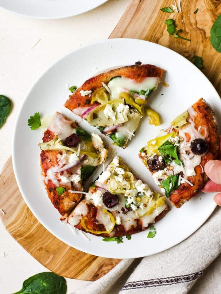 Greek pita pizza slices on white plate with hand picking up a slice.
