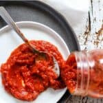 Roasted red pepper pizza sauce poured out from glass jar onto white plate with spoon.