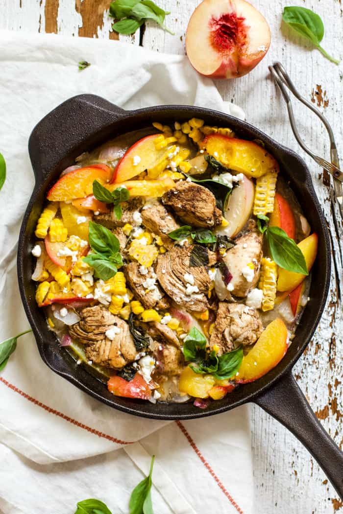 Peach basil balsamic chicken in a cast iron skillet with fresh basil and goat cheese, on top of a white linen napkin on a white farmhouse-style surface