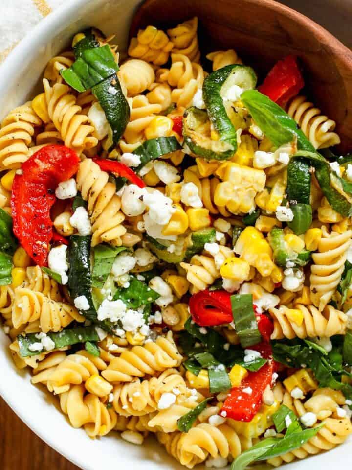 Late summer veggie pasta salad with roasted veggies and goat cheese in white bowl with wooden spoon.