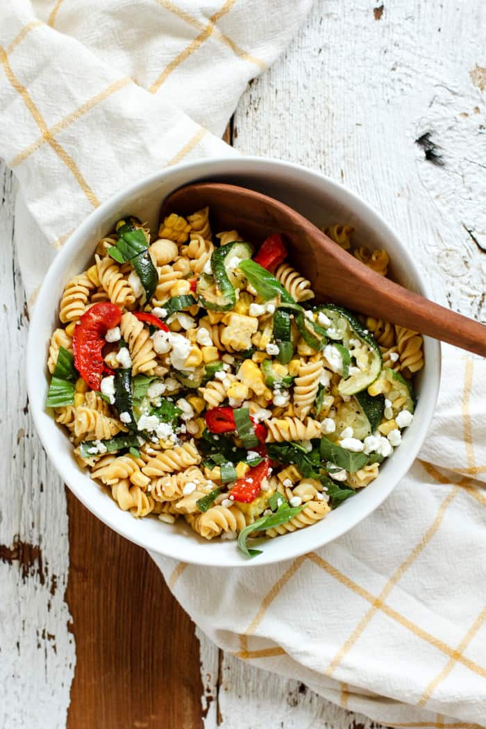 Late summer veggie pasta salad with red peppers, corn, zucchini, fresh basil, and goat cheese, in a white bowl with a wooden spoon