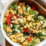 Late summer veggie pasta salad with red peppers, corn, zucchini, fresh basil, and goat cheese, in a white bowl with a wooden spoon