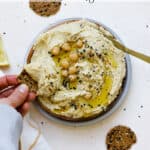 Everything bagel hummus served on wooden plate, drizzled with olive oil, with round seedy crackers on the side and being dipped.