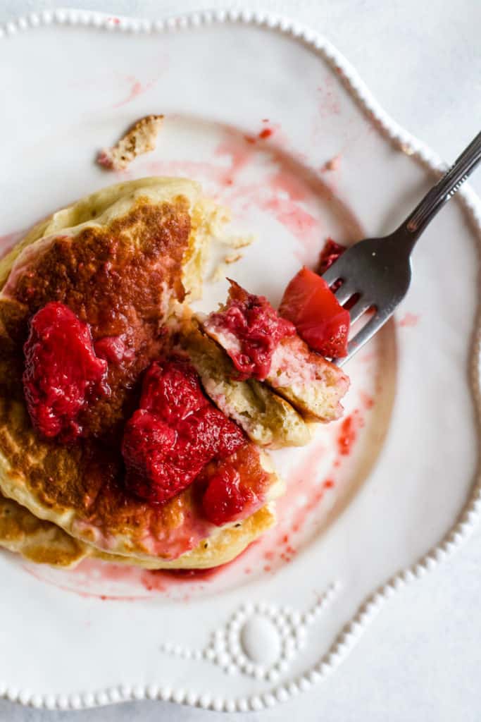 Stack of whole wheat Greek yogurt pancakes with strawberry compote on top, on white plate with fork with bite of pancake on it.