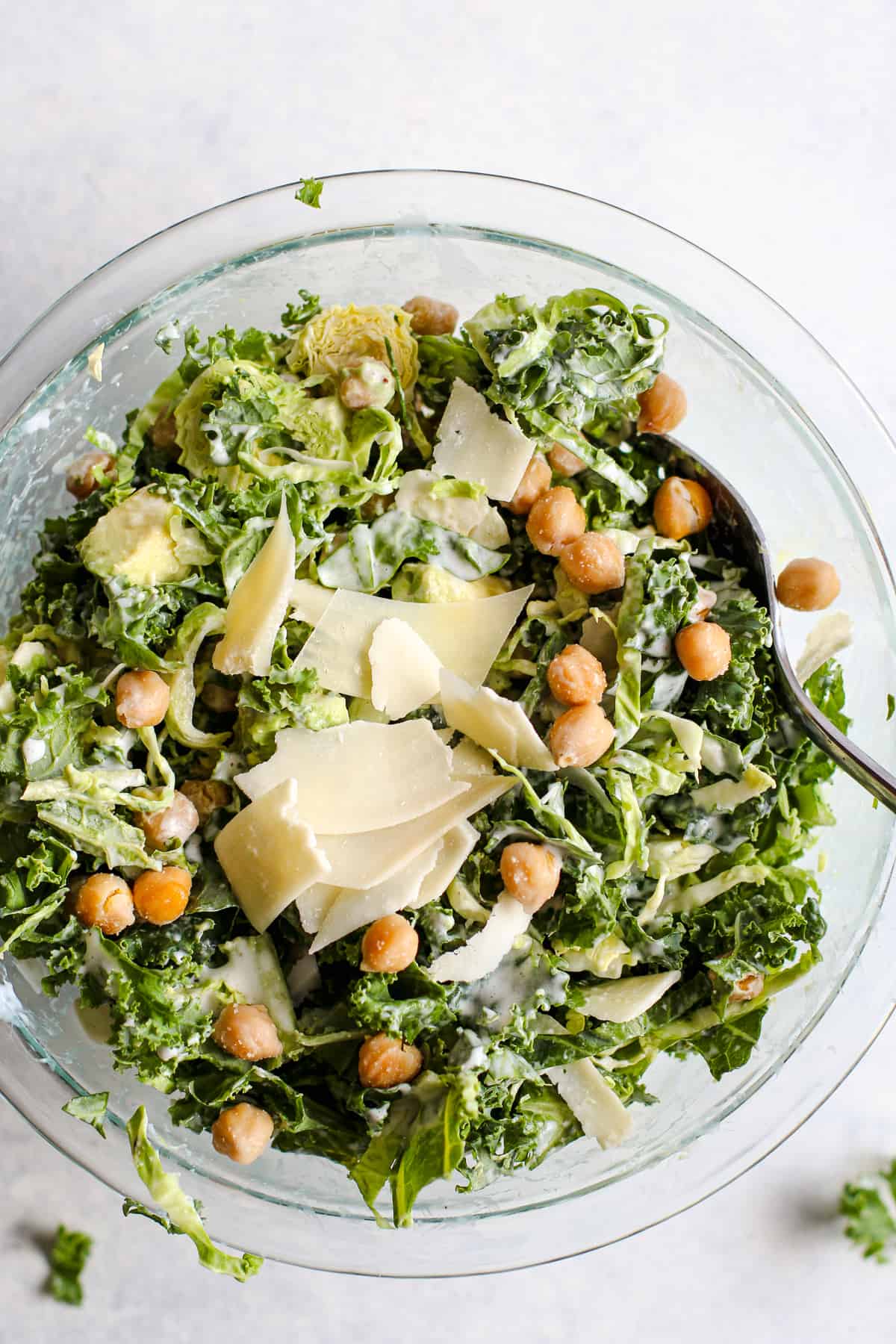 Kale caesar salad with garlic roasted chickpeas and thinly sliced parmesan in a clear glass bowl. A serving spoon rests in the bowl and the bowl sits on a light blue and white surface.