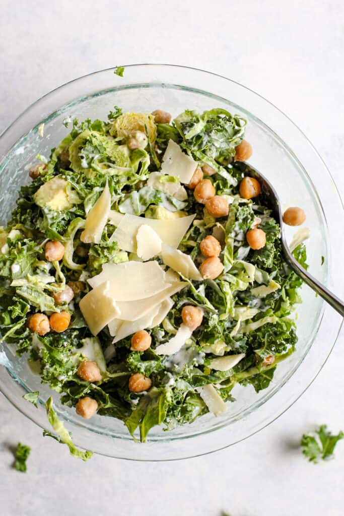 Kale caesar salad with garlic roasted chickpeas and thinly sliced parmesan in a clear glass bowl. A serving spoon rests in the bowl and the bowl sits on a light blue and white surface.