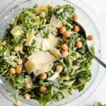 Kale caesar salad with garlic roasted chickpeas and thinly sliced parmesan in a clear glass bowl. A serving spoon rests in the bowl and the bowl sits on a light blue and white surface.