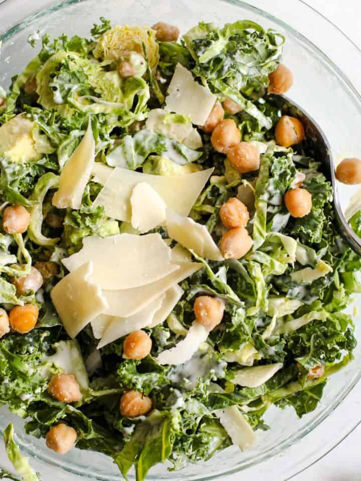 Kale caesar salad with garlic roasted chickpeas and thinly sliced parmesan in a clear glass bowl. A serving spoon rests in the bowl and the bowl sits on a light blue and white surface.