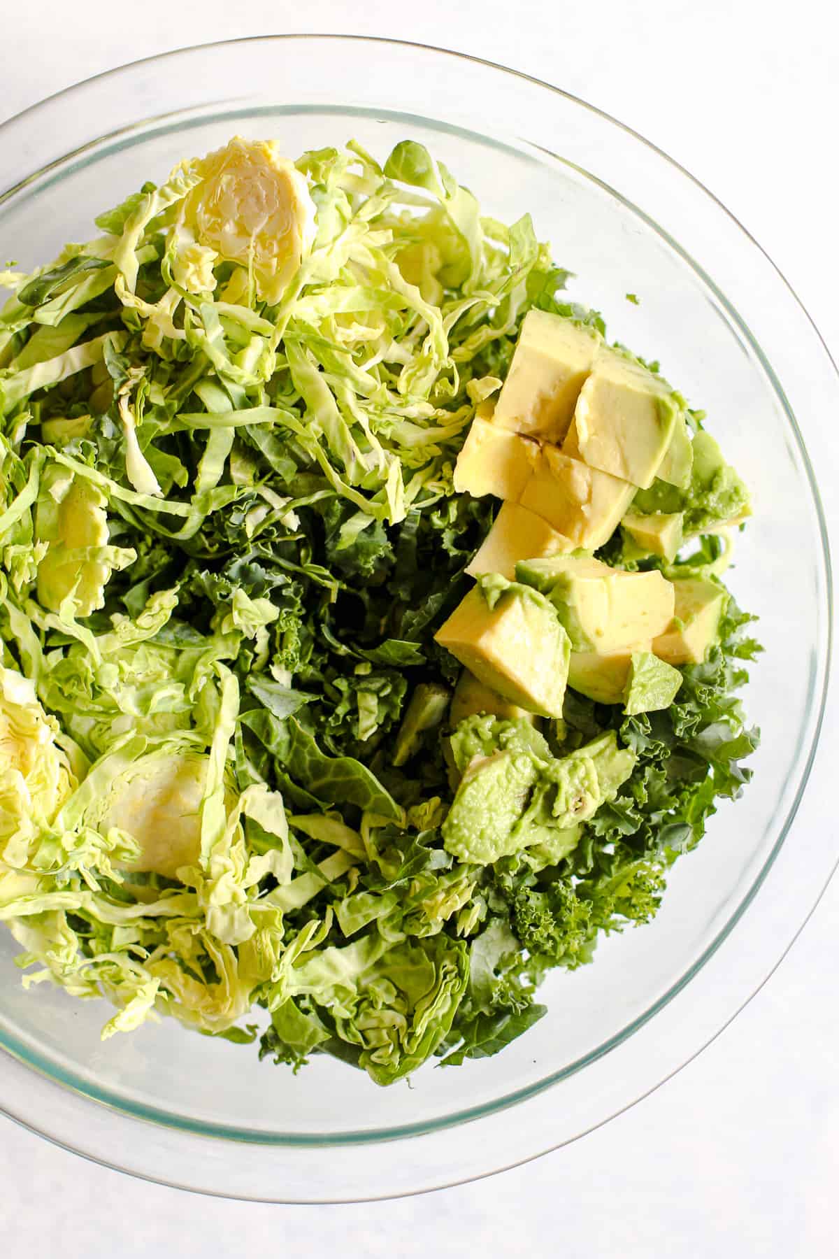 Curly kale, thinly shaved Brussels sprouts, and diced avocado are in a clear glass bowl on a light blue and white surface.