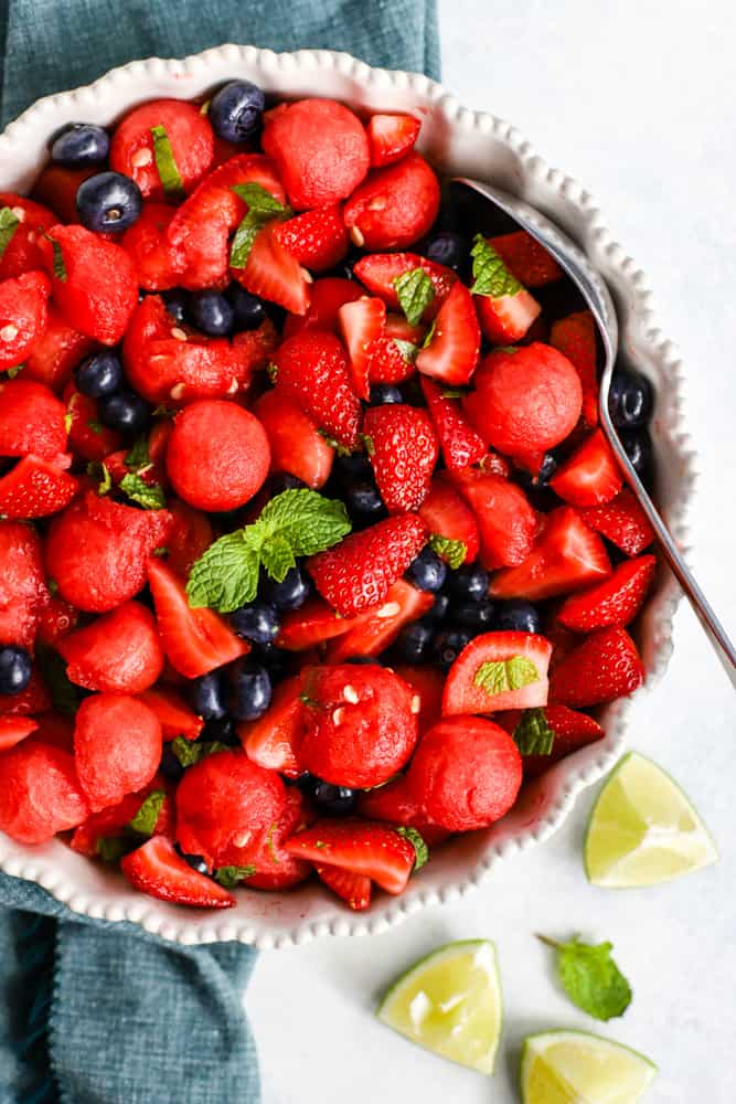 Honey lime mint watermelon berry salad in large with serving dish on light surface with blue linen, garnished with limes and mint