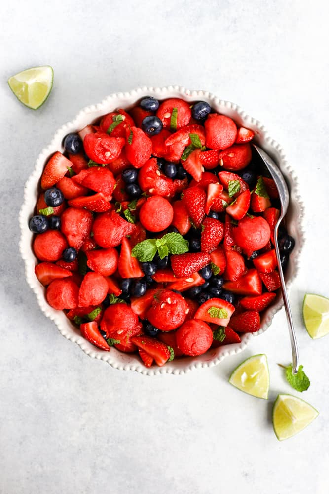 Honey lime mint watermelon berry salad in large with serving dish on light surface garnished with limes and mint