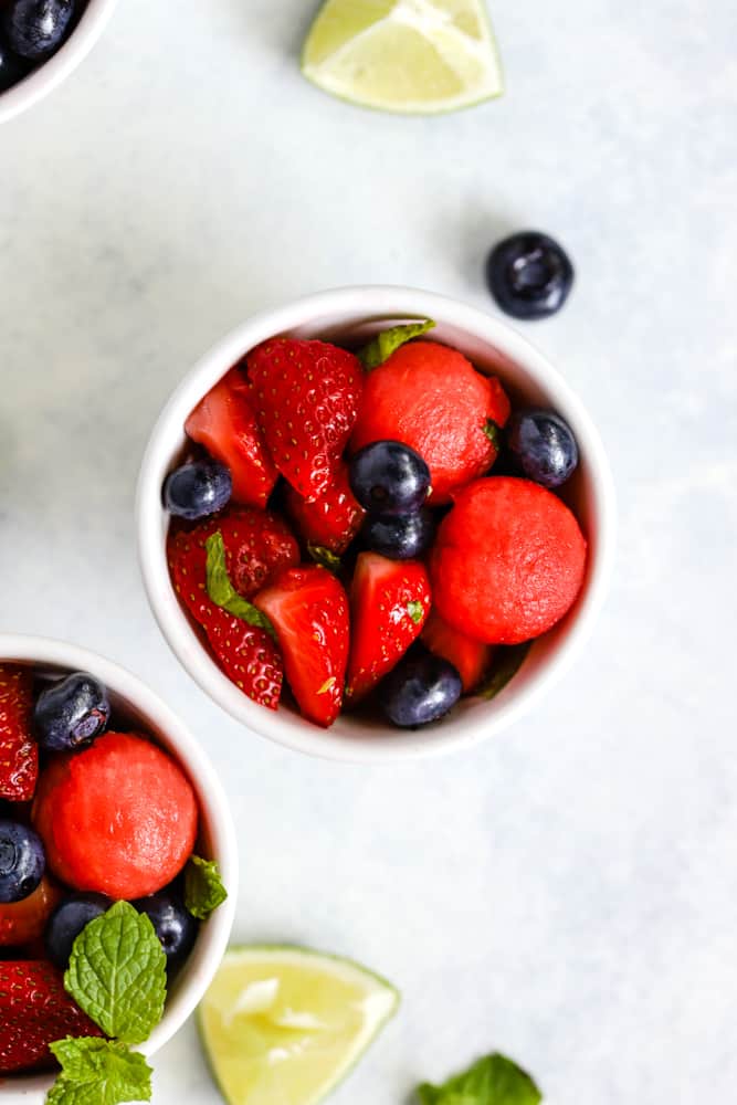 Honey lime mint watermelon berry salad in small white bowls on light surface with lime and blueberry