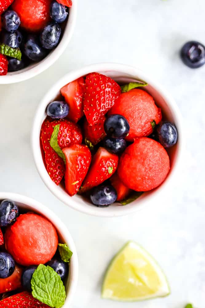 Honey lime mint watermelon berry salad in small white bowls on light surface with lime and blueberry