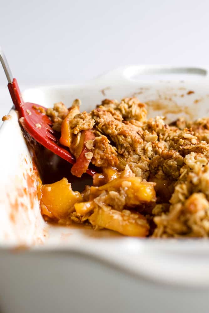 Baked healthy peach crisp in white ceramic baking dish with red spatula