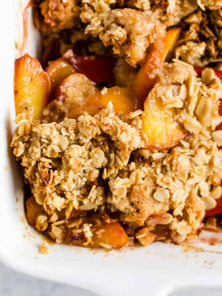 Healthy peach crisp in white ceramic baking dish with red spatula scooping out a square piece