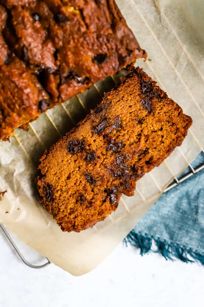 Slice of healthy pumpkin chocolate chip bread next to loaf, on parchment over cooling rack