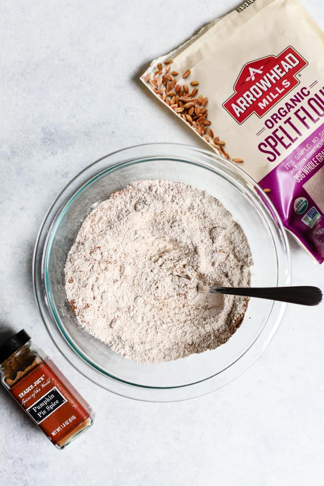 Flour, pumpkin pie spice, baking soda, and salt in clear bowl, with spelt flour bag and pumpkin pie spice jar on the side