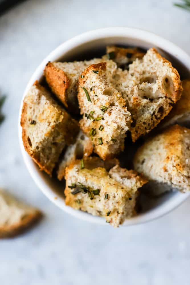 Baked homemade whole wheat croutons in small white bowl with rosemary sprigs in background