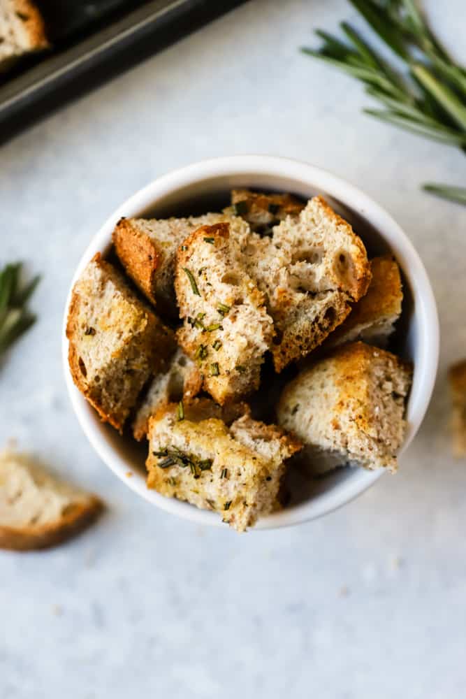 Baked homemade whole wheat croutons in small white bowl with rosemary sprigs in background