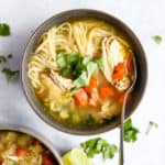 Ginger lime chickek noodle soup in dark gray bowl garnished with cilantro, on light blue surface with teal linen