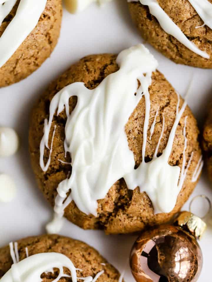 Flourless almond butter cookies drizzled with white chocolate, on white marble surface with small copper ornament and white chocolate chips sprinkled around