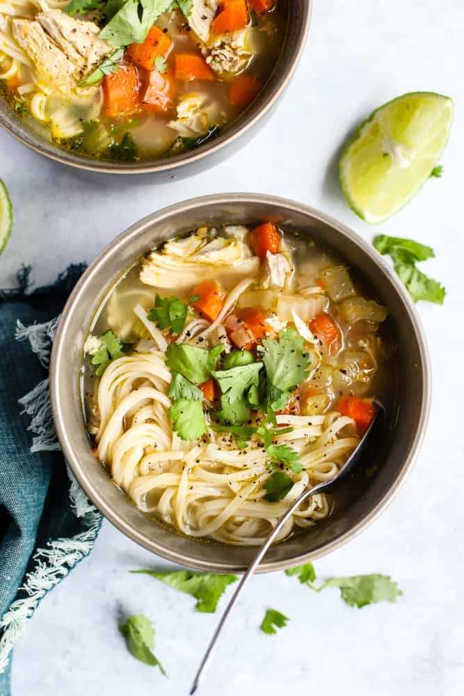 Ginger lime chickek noodle soup in dark gray bowl garnished with cilantro, on light blue surface with teal linen