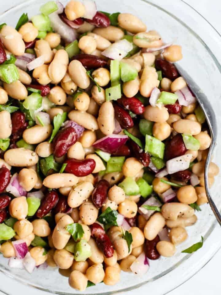 Modern three bean salad in clear glass bowl with spoon, on light blue and white surface.