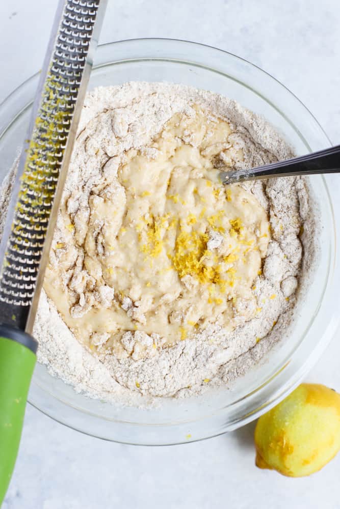 Folding flour mixture into wet mixture, showing lemon zest before folding in, in a clear bowl next to partially zested lemon and zester