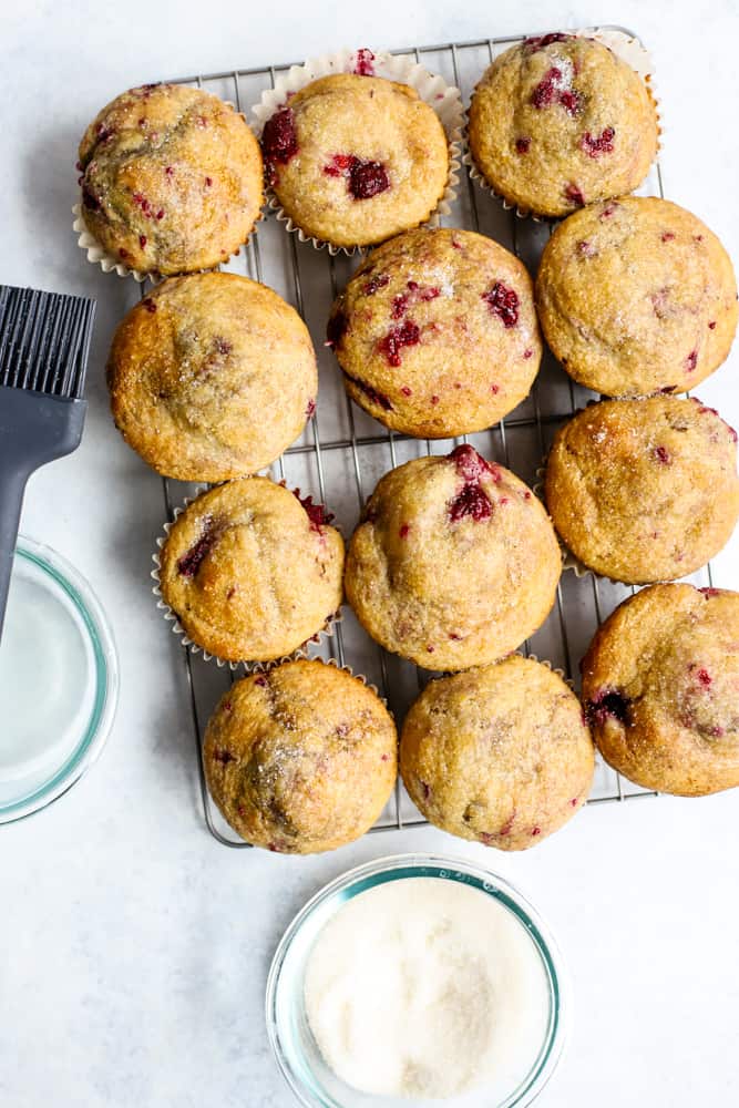 Baked raspberry lemon muffins on cooling rack next to glass bowls with coconut oil and sugar, plus silicone brush