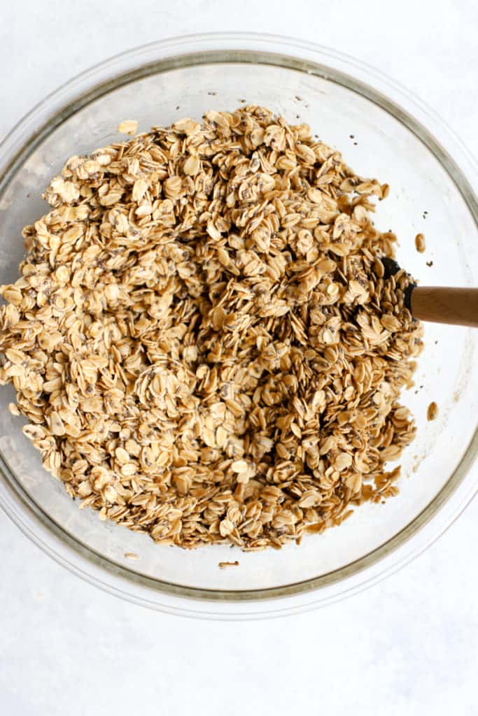 Oats, flour, chia seeds, cinnamon, and salt in a clear glass bowl, stirred together with maple syrup, coconut oil, and vanilla extract