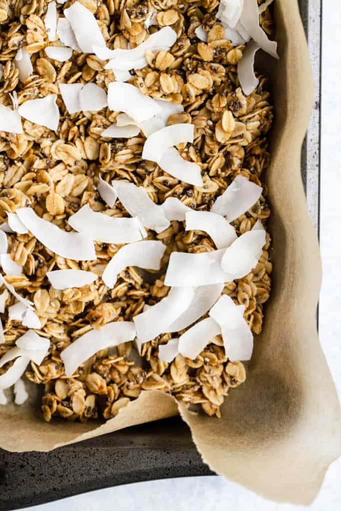 Simple coconut chia granola on parchment paper lined sheet pan with coconut just added before putting back in oven to toast