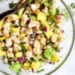 Shrimp mango and avocado salad in clear bowl with copper serving spoon set on light blue and white background