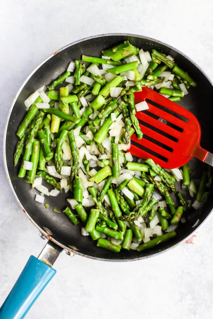 Asparagus pieces and diced onion in sauté pan with red spatula.