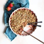 Healthy strawberry crisp in cream-colored serving dish, with pieces missing, copper serving spoon, and two small forks in the dish; next to teal linen with two fresh strawberries on the side