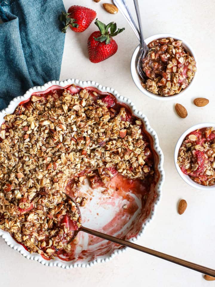 Healthy strawberry crisp in cream-colored pie dish with copper serving spoon and two small white bowls with more crisp, with almonds and fresh strawberries on the side