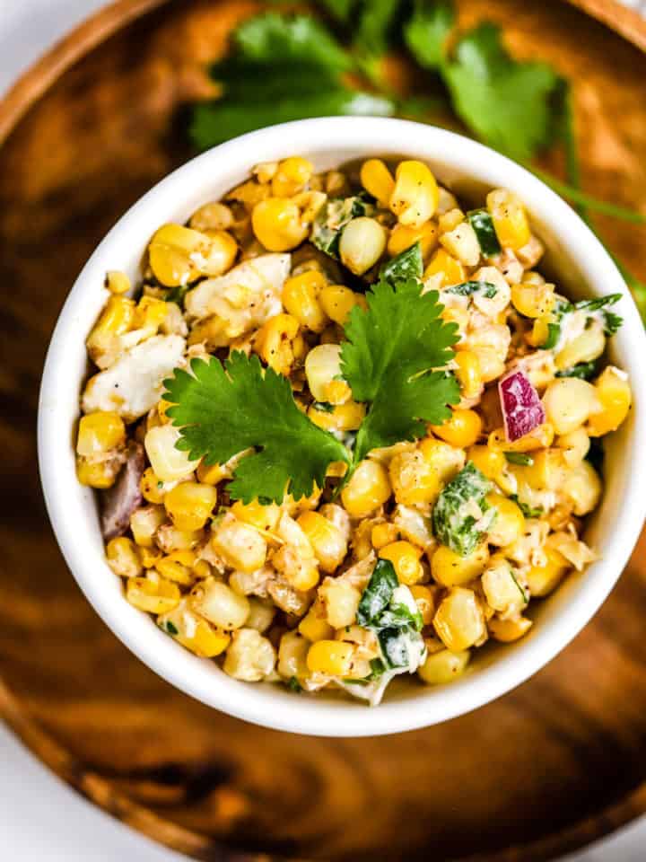 Healthy Mexican street corn salad in white bowl on wooden plate with fresh cilantro garnish