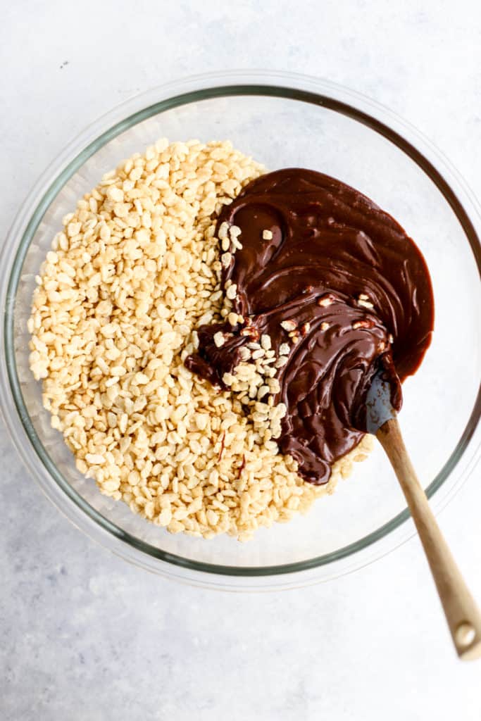 Date syrup and peanut butter mixture poured into clear glass bowl with brown rice crisps with spatula, not yet stirred together