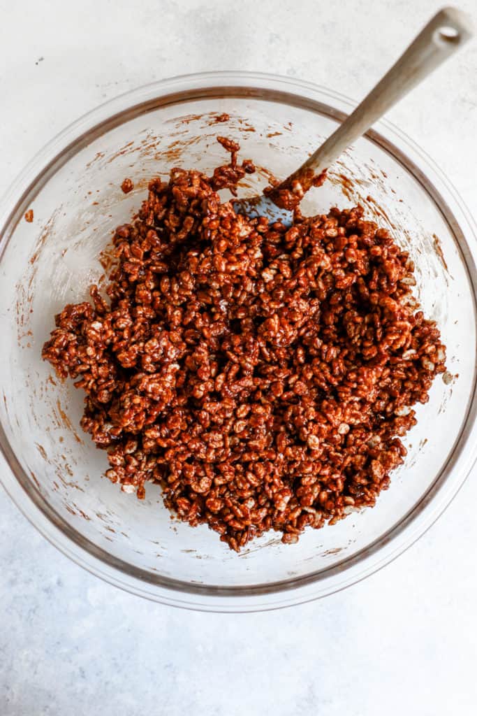 Date syrup and peanut butter mixture fully combined with brown rice crisps in clear glass bowl with spatula