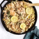 One skillet chicken with leeks and farro in cast iron skillet fresh out of the oven with lemon slices on top, wooden spoon, teal linen around the handle, on white and blue surface