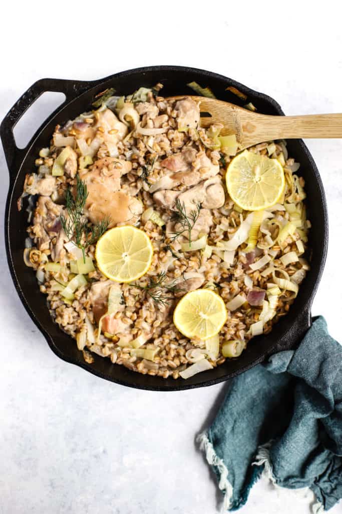 One skillet chicken with leeks and farro in cast iron skillet fresh out of the oven with lemon slices on top, wooden spoon, teal linen around the handle, on white and blue surface